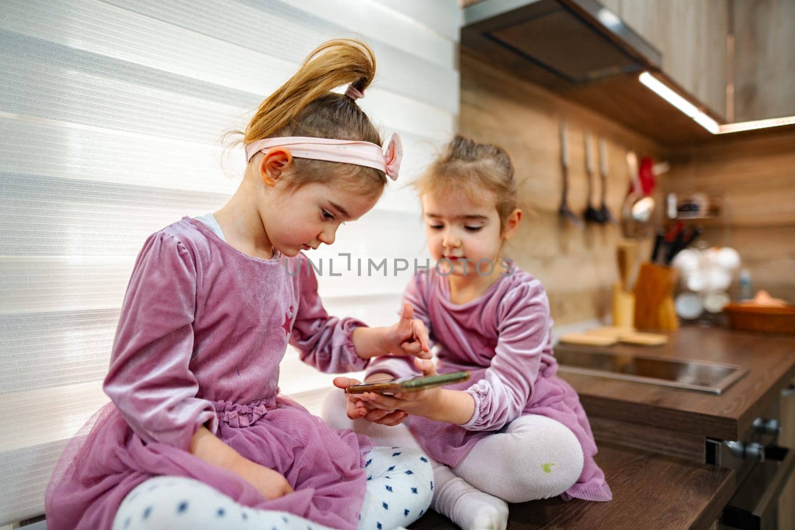 In a cozy, well-lit kitchen, two little girls in matching pink dresses enjoy sharing a tablet, engrossed in their activity while playfully interacting with each other and their surroundings.