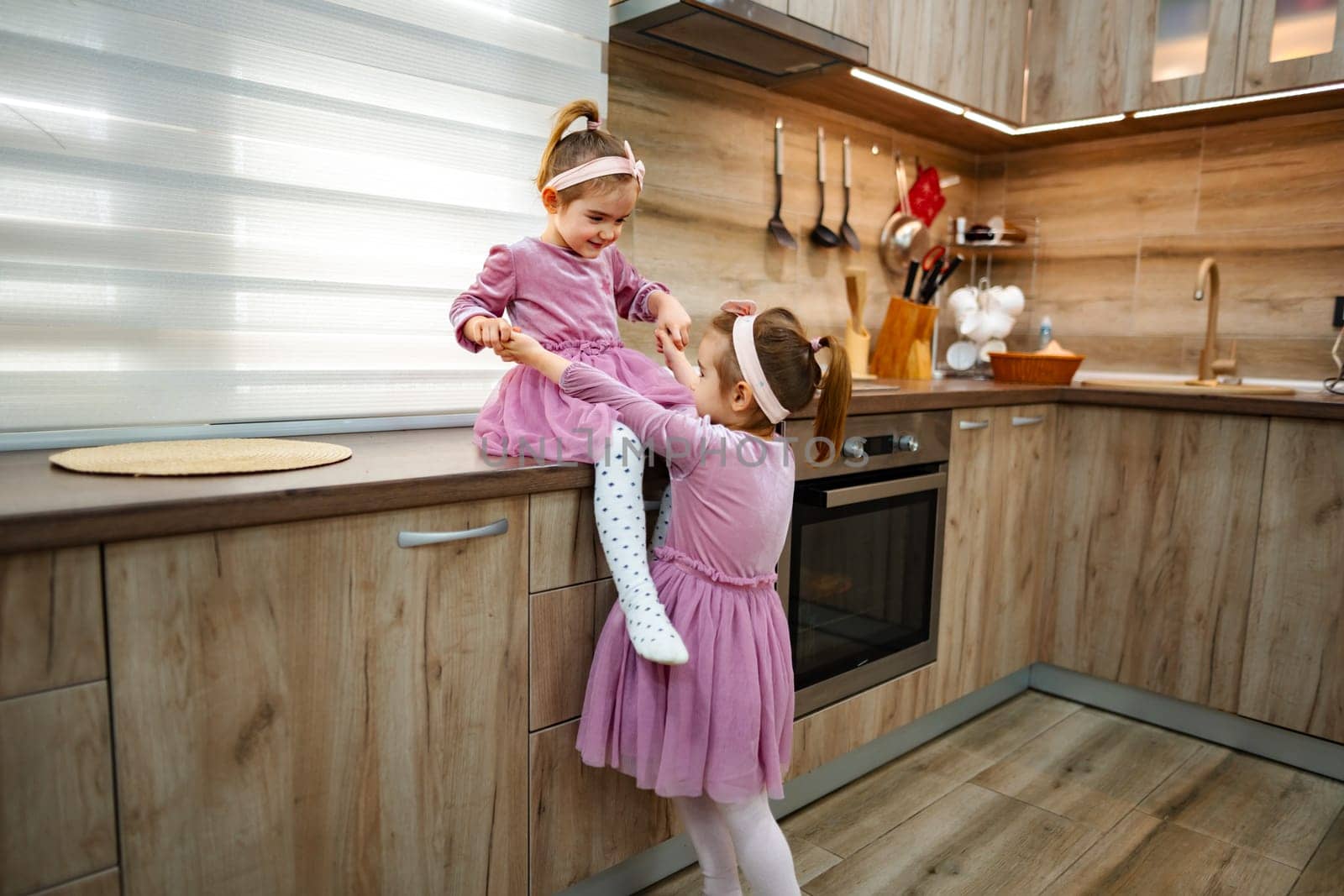 In a cozy and stylish kitchen, two young girls are immersed in playful fun, one perched on the counter while the other reaches up joyfully, fostering creativity and laughter in their enchanting world.