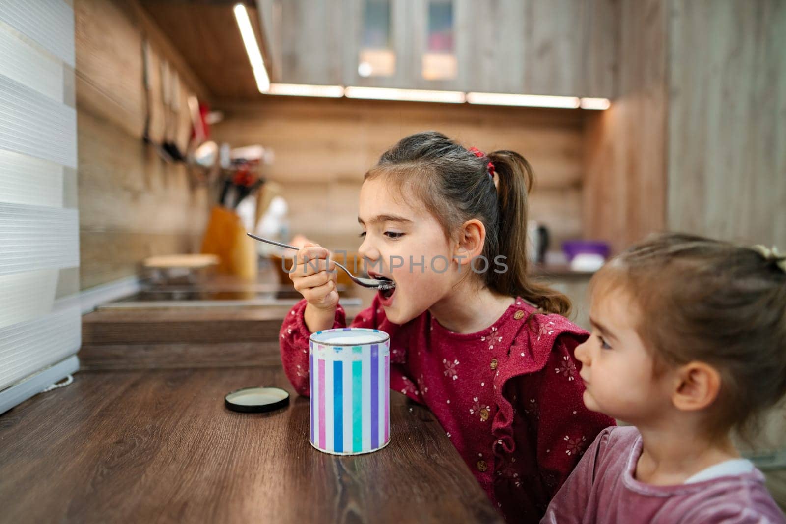 In a warm, inviting kitchen, two sisters share a delightful moment as one eagerly tastes a treat from a colorful container. The atmosphere is filled with joy and laughter, highlighting their bond.