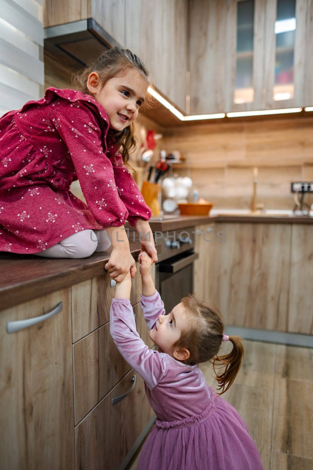 In a warm and inviting kitchen, two young sisters engage in a delightful game. One sister, dressed in a vibrant dress, is perched on the counter as the younger one, eager, reaches up to assist her.