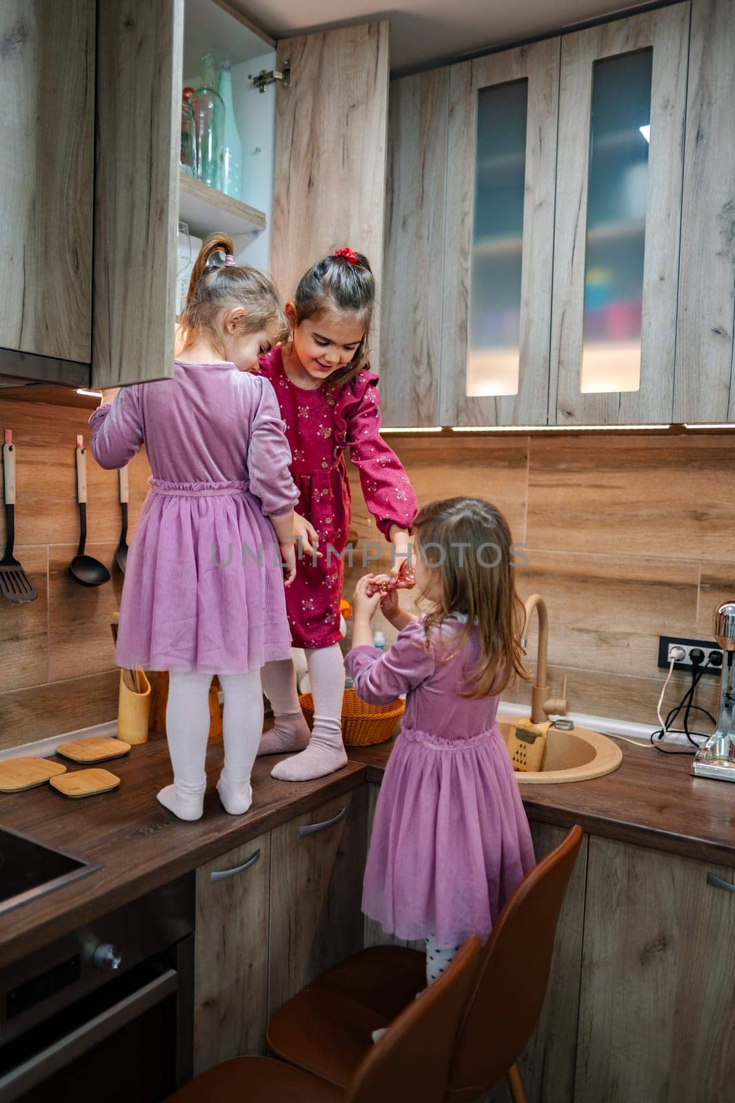 Three young girls, dressed in matching purple dresses, playfully explore a modern kitchen, reaching for ingredients as they prepare a snack, creating joyful laughter and cherished memories.