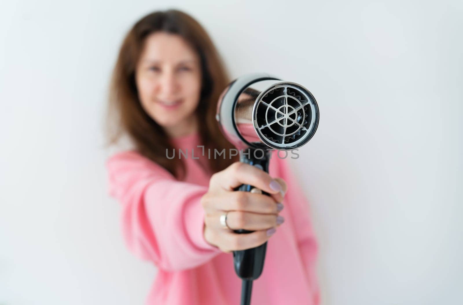 A smiling woman in a pink sweater holds out a hairdryer, ready to dry her hair.