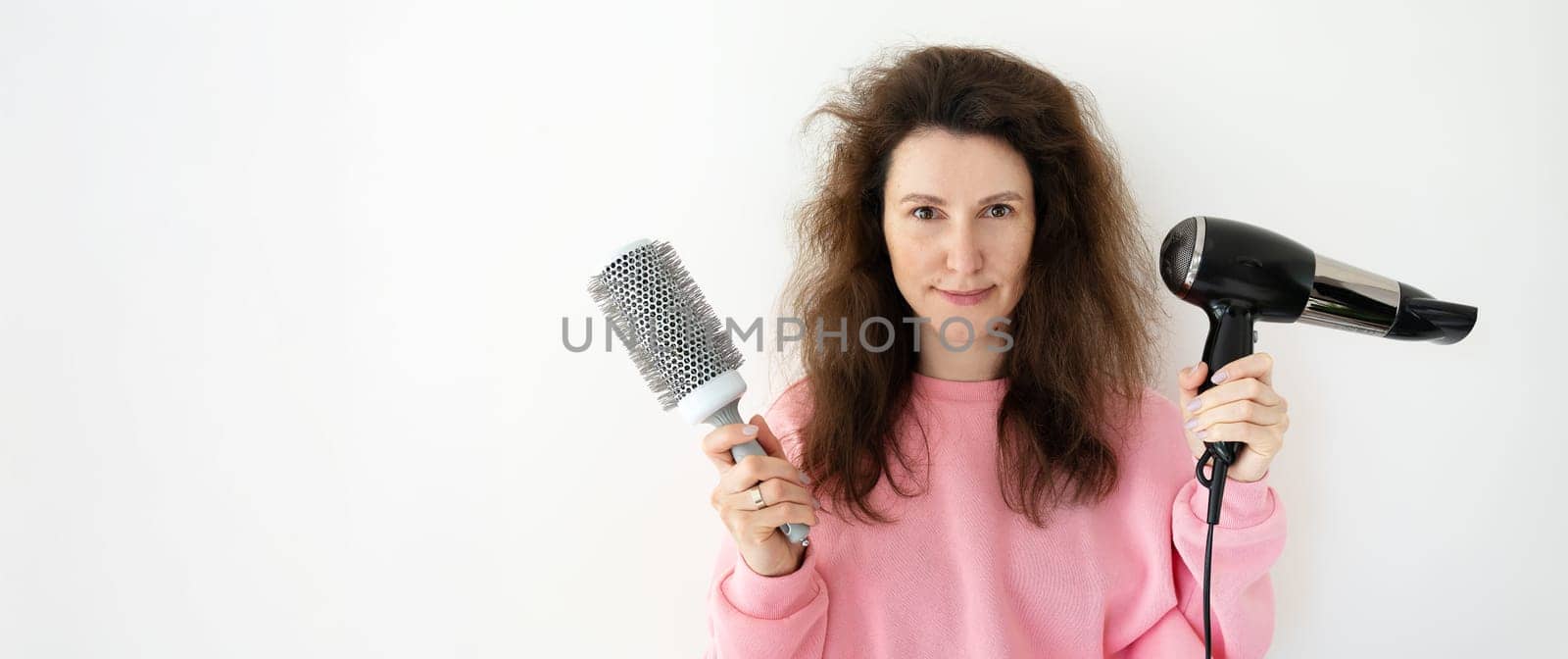 A woman with messy hair holds a round hairbrush and hairdryer, preparing to style her hair.