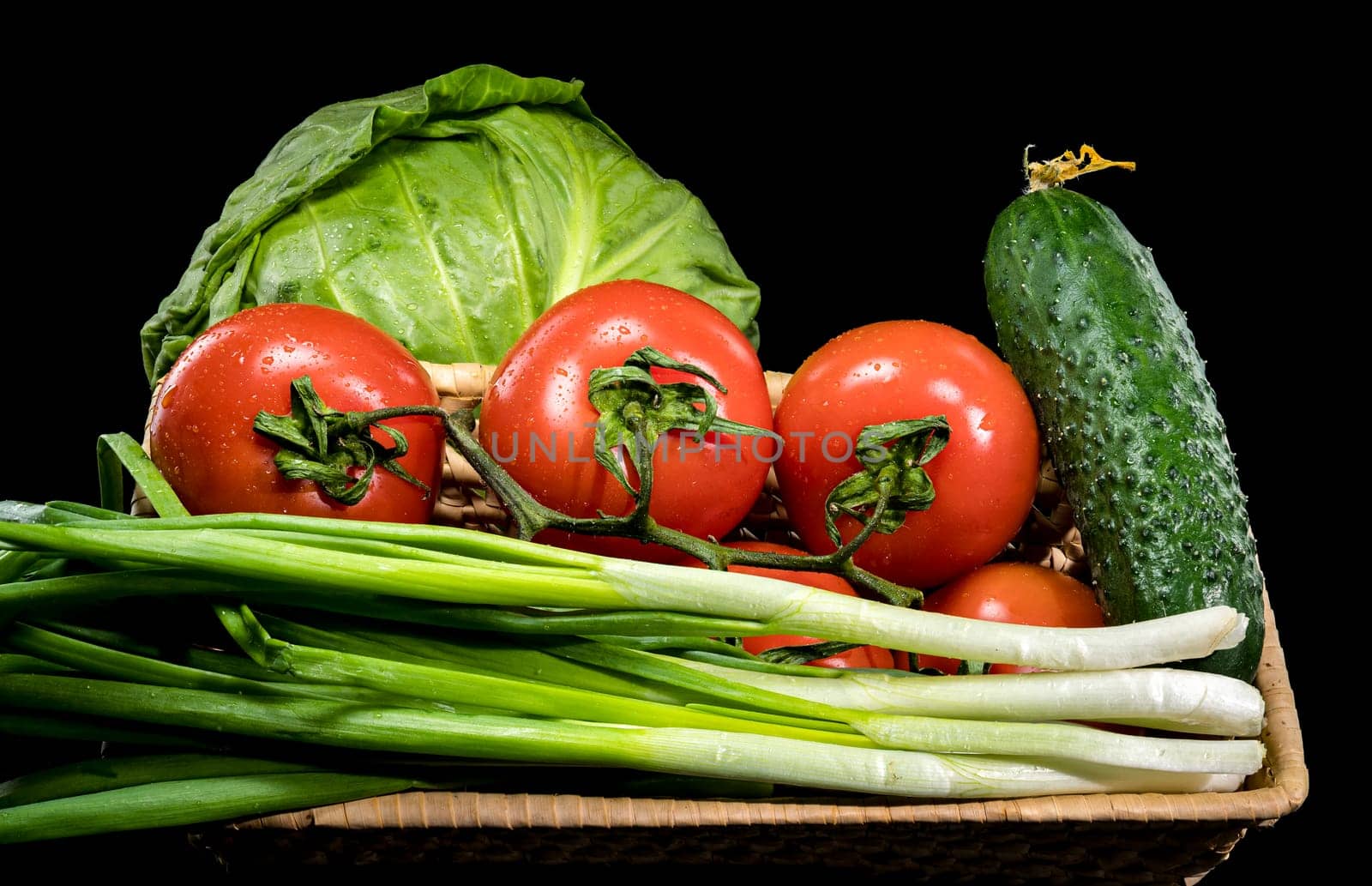 Fresh green cabbage, ripe red tomatoes, cucumber, and spring onions arranged on a wooden tray against a black background 