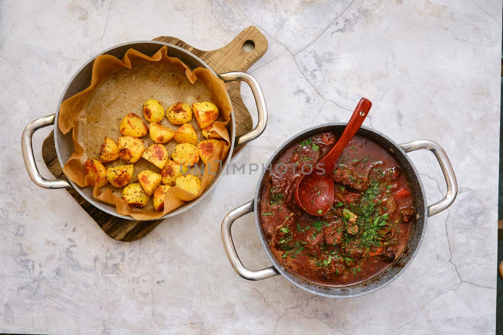 A hearty beef stew in a pot, topped with fresh herbs. Beside it, golden roasted potatoes are presented in a baking pan, showcasing a cozy meal ready to be enjoyed in a warm kitchen.