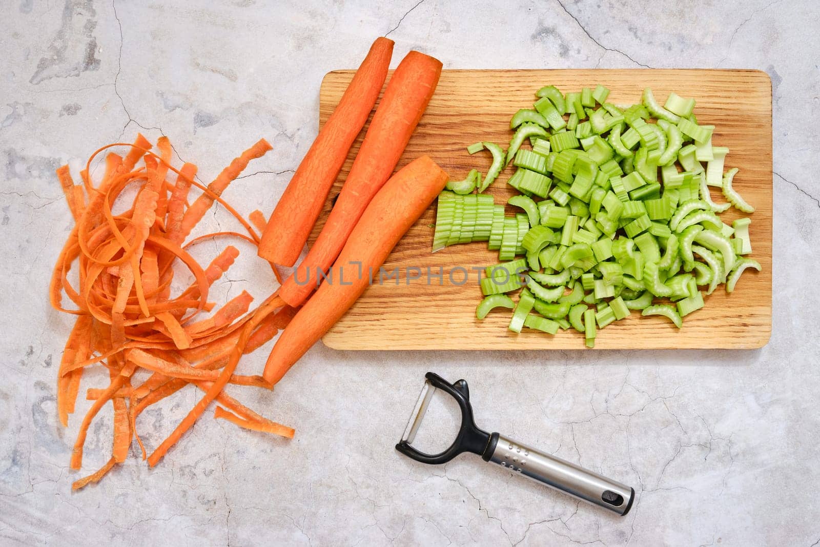 Peeled carrots sit alongside finely chopped green celery on a wooden cutting board. Vegetable peels are scattered around, indicating preparation for a healthy meal in a modern kitchen.