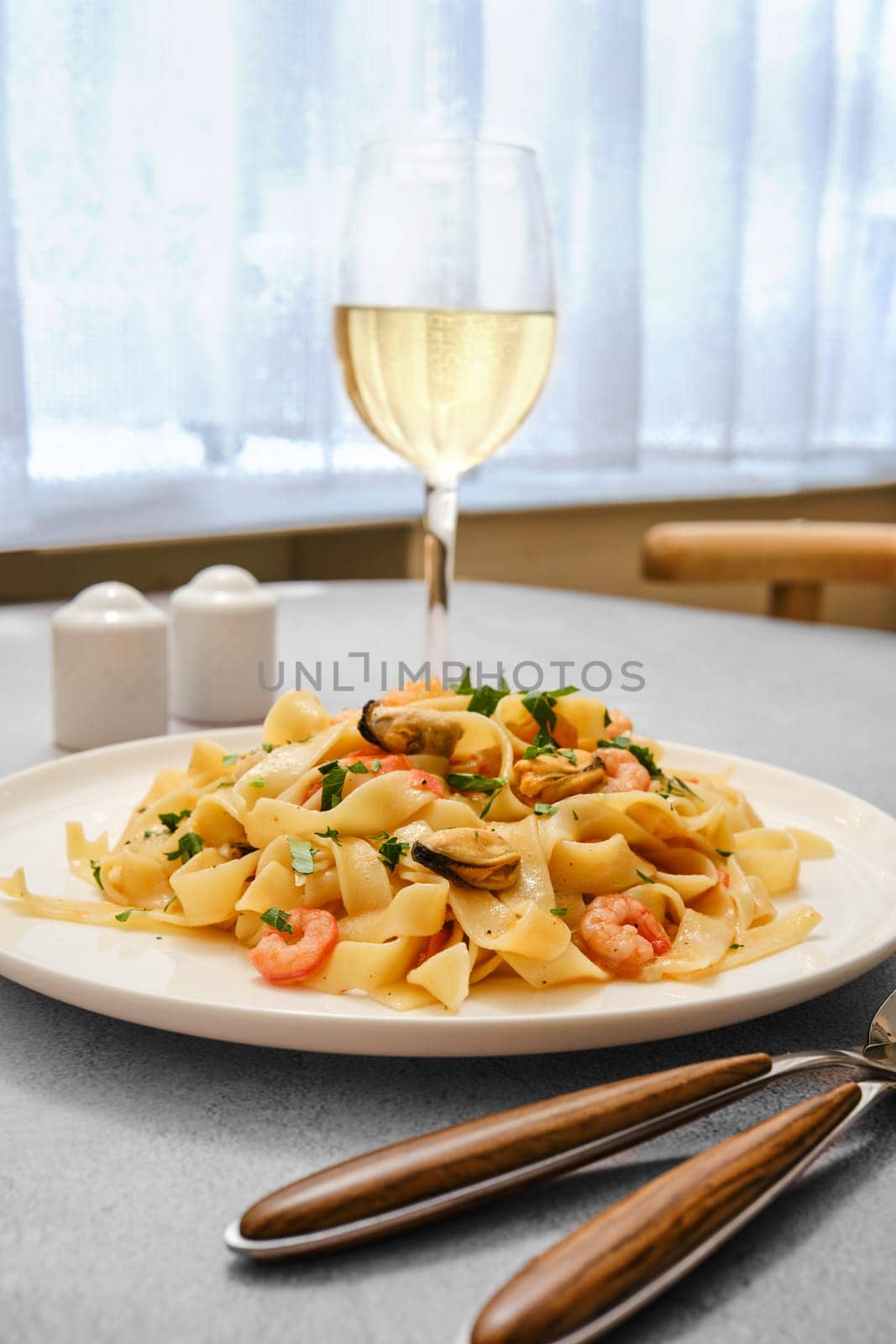 A plate of seafood pasta with shrimp and clams on a kitchen table. A glass of white wine stands nearby, complementing the meal in a bright and inviting dining space during midday.