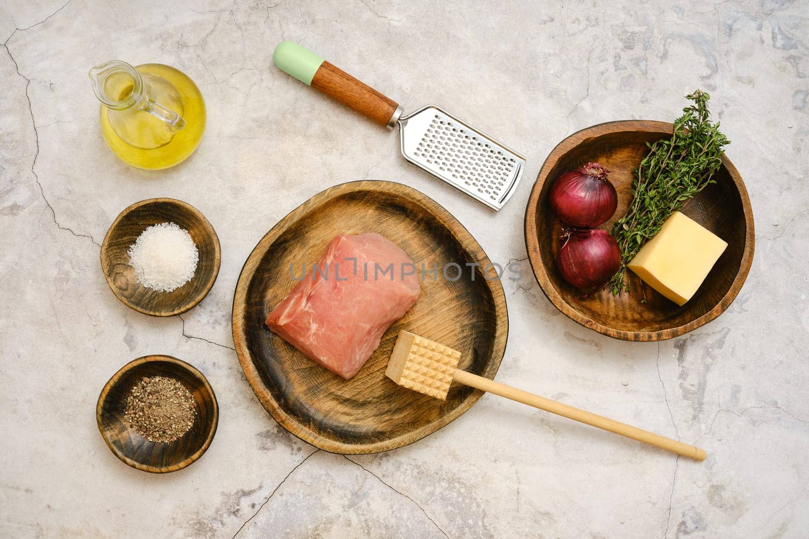 Freshly sourced pork chop is laid on a wooden plate alongside red onions, thyme, cheese, and seasoning. Cooking tools like a grater and a wooden mallet are nearby, ready for meal prep.