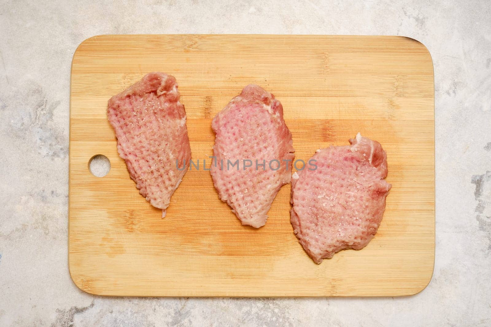 Three raw pork chop cuts are on a wooden chopping board. The fresh meat is awaiting seasoning before being cooked on a stovetop or grill for a delicious meal.