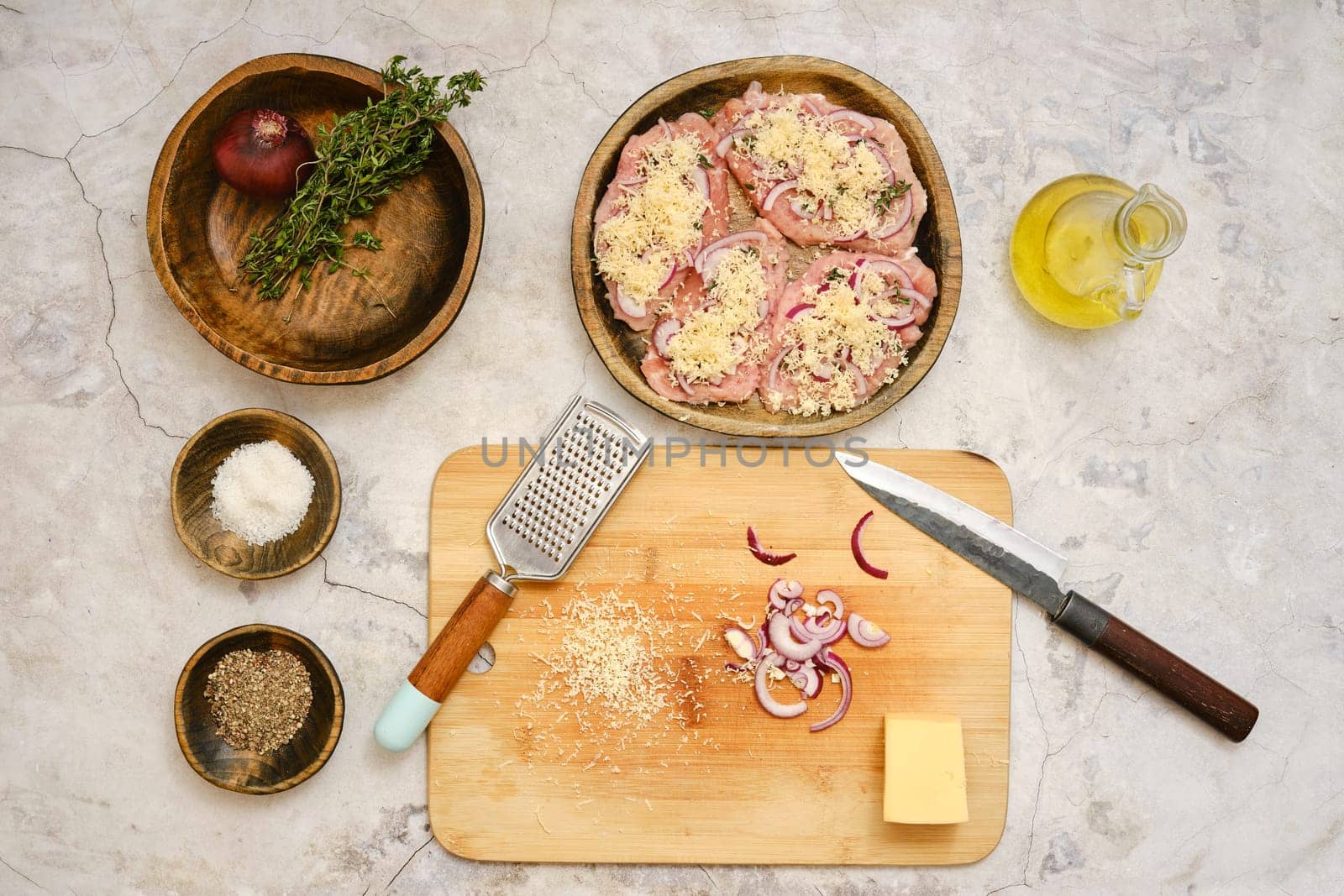 Fresh pork chops are topped with onion and grated cheese on a wooden plate. Ingredients like salt, pepper, and cheese block are ready for seasoning and cooking.