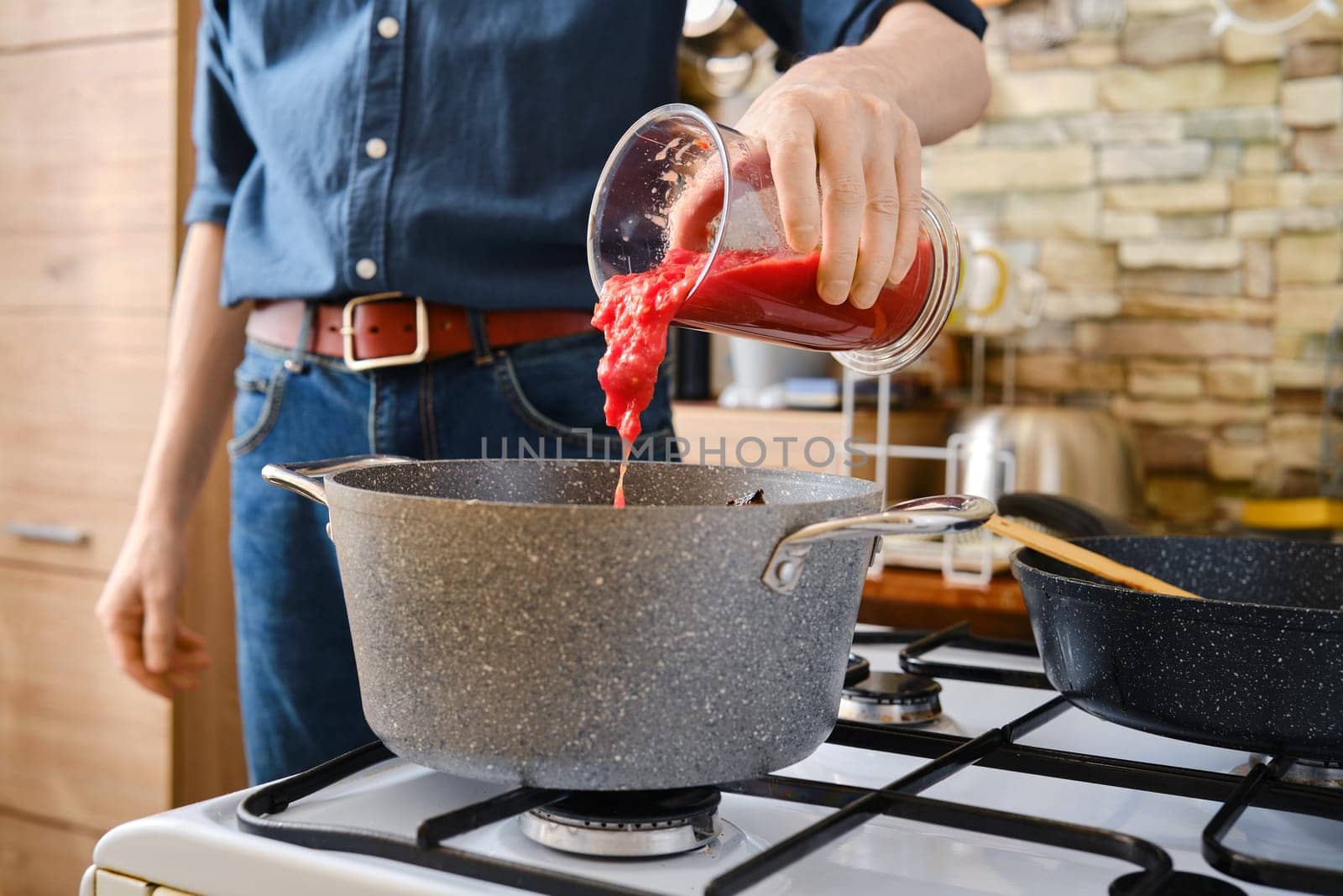 A person pours tomato sauce from a jar into a pot on the stove. The kitchen features modern decor with a warm ambiance.