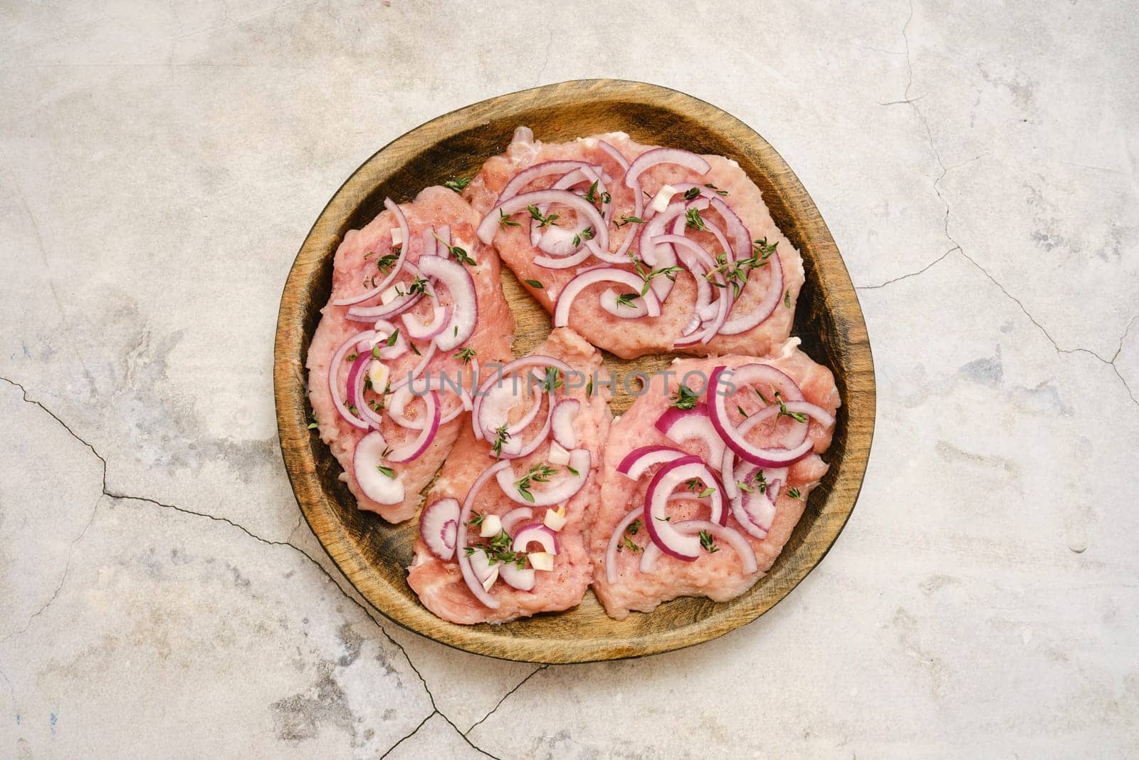 Four pork chops are on a wooden plate, topped with thinly sliced onions and fresh herbs. This dish looks ready for grilling or baking, promising a flavorful meal.