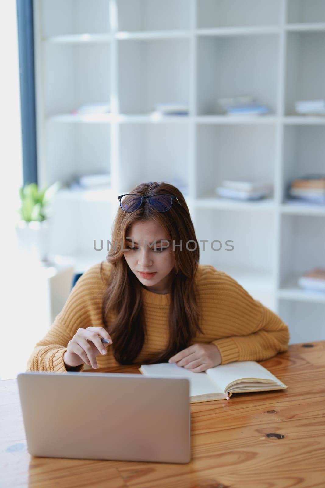 Smiling young woman engaged in a joyful online e-learning video call, gesturing while holding a book with headphones on a wooden table nearby.
