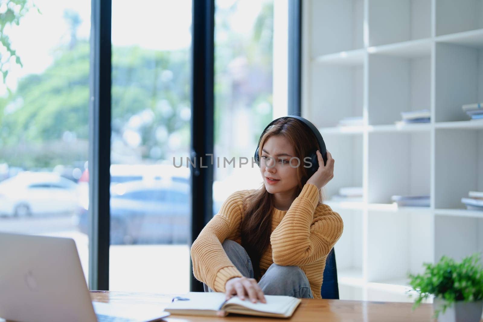 Young woman enjoying a relaxing music break at her desk while using headphones, writing in a notebook, and working on her laptop for calm inspiration.