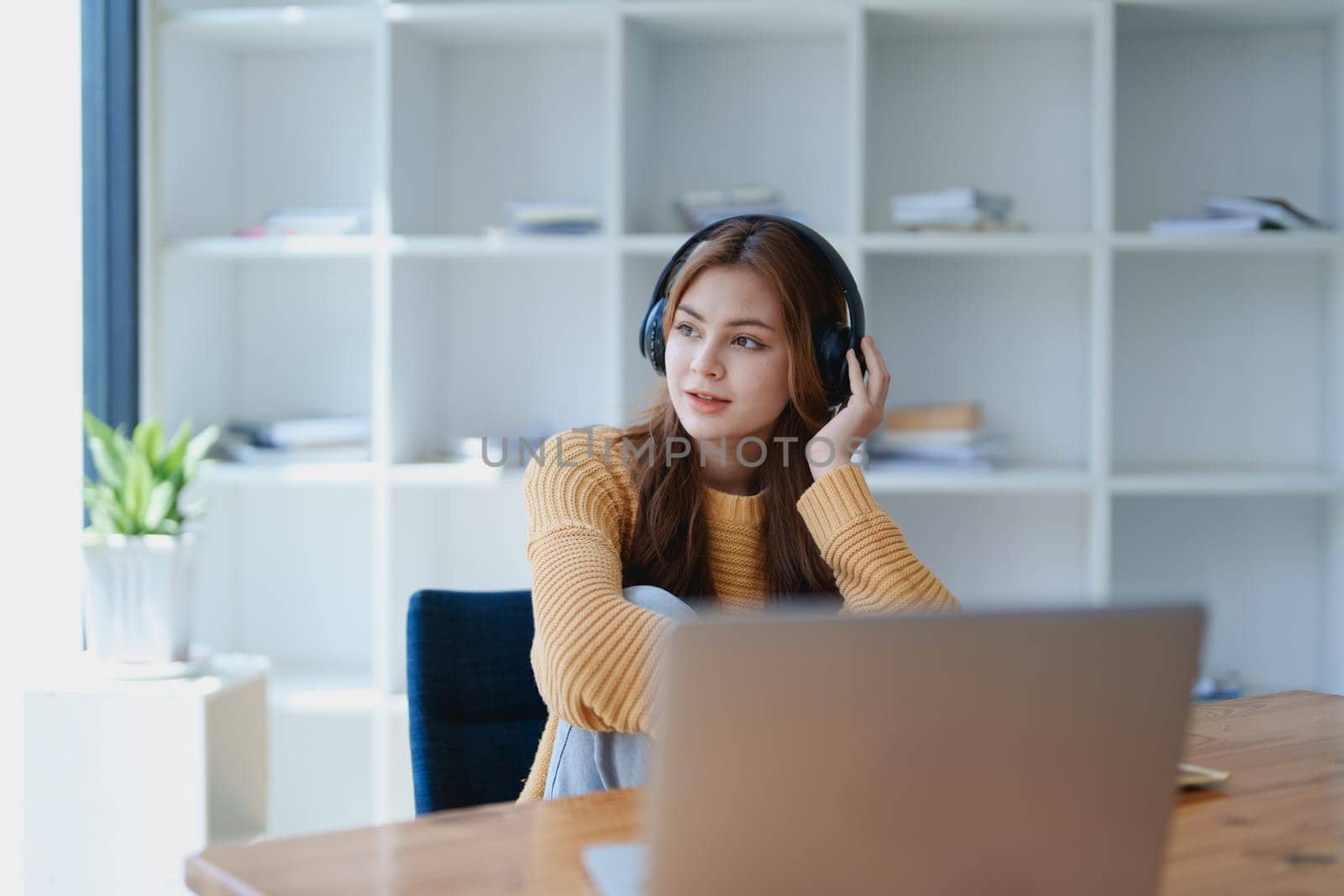 Young woman enjoying a relaxing music break at her desk while using headphones, writing in a notebook, and working on her laptop for calm inspiration.