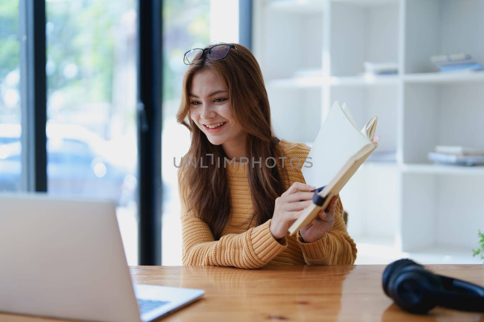 Smiling young woman engaged in a joyful online e-learning video call, gesturing while holding a book with headphones on a wooden table nearby.