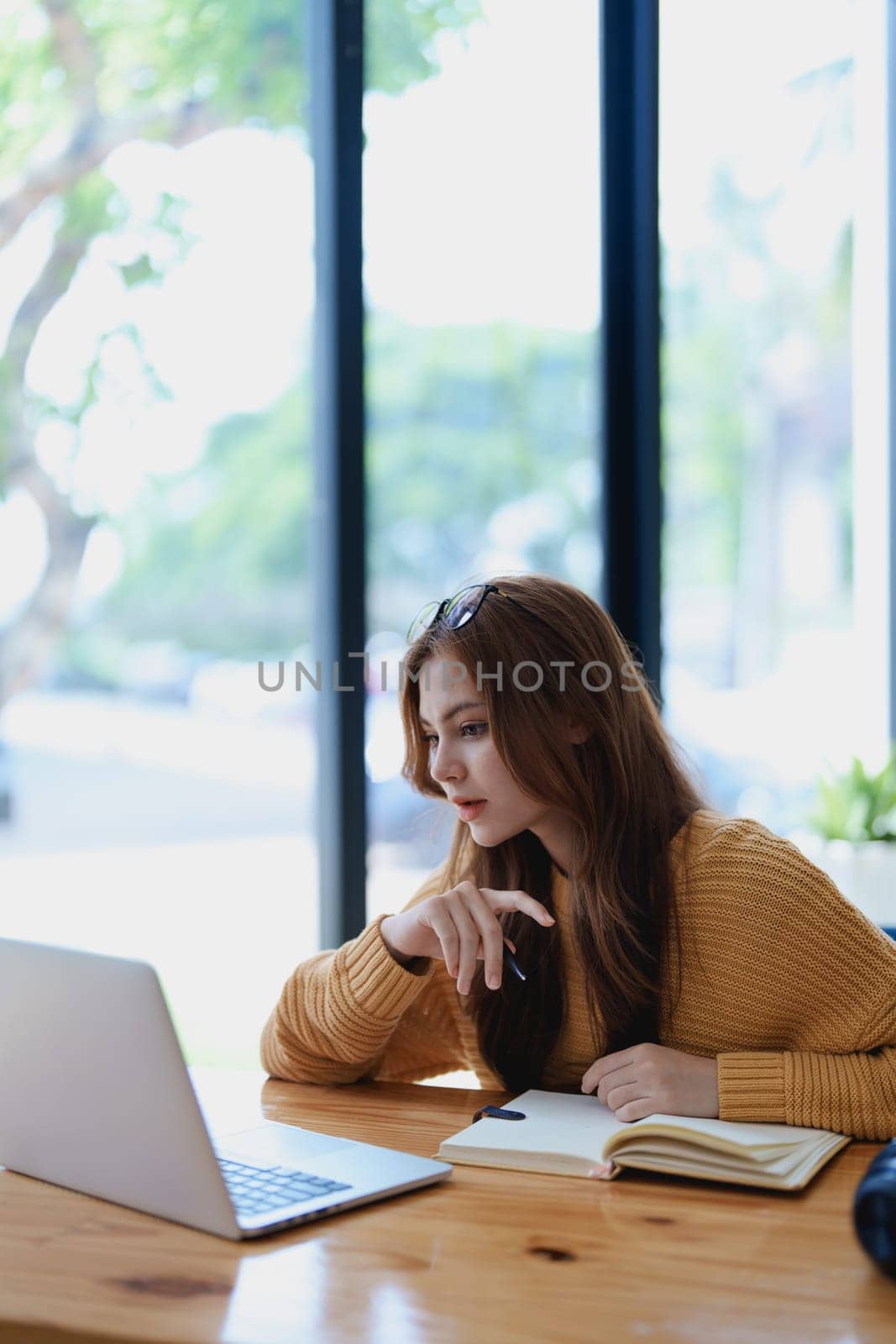 Smiling young woman engaged in a joyful online e-learning video call, gesturing while holding a book with headphones on a wooden table nearby.
