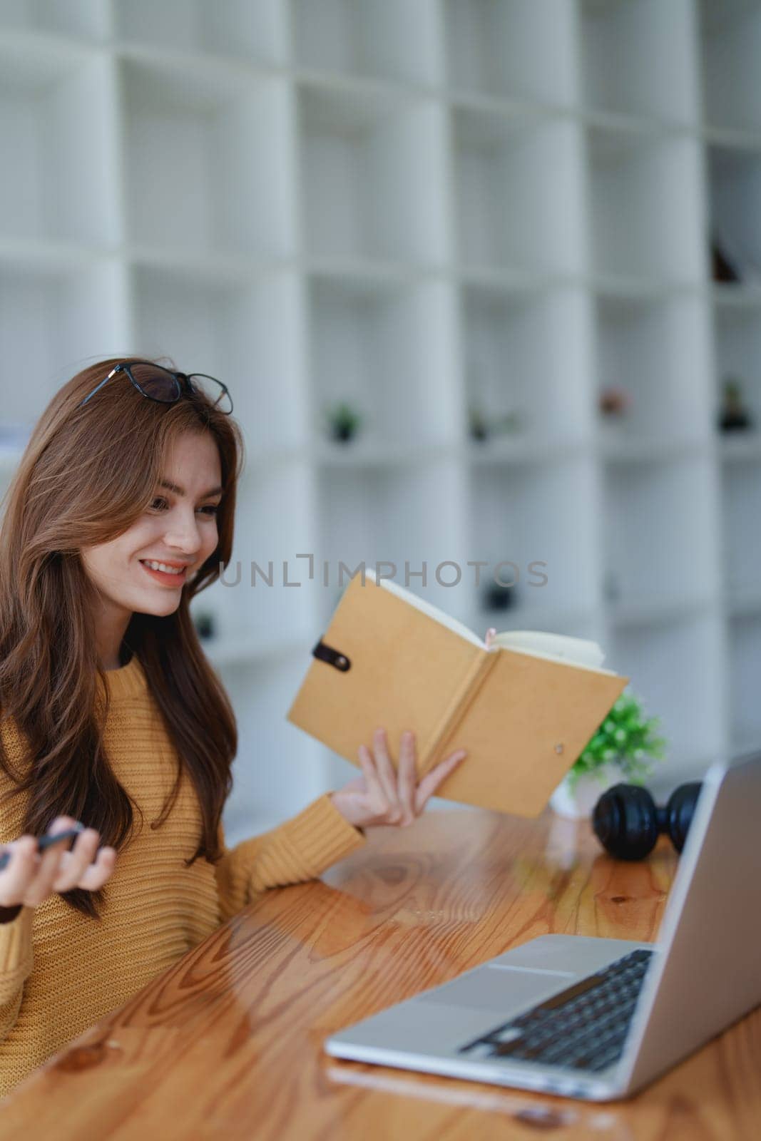 Smiling young woman engaged in a joyful online e-learning video call, gesturing while holding a book with headphones on a wooden table nearby.
