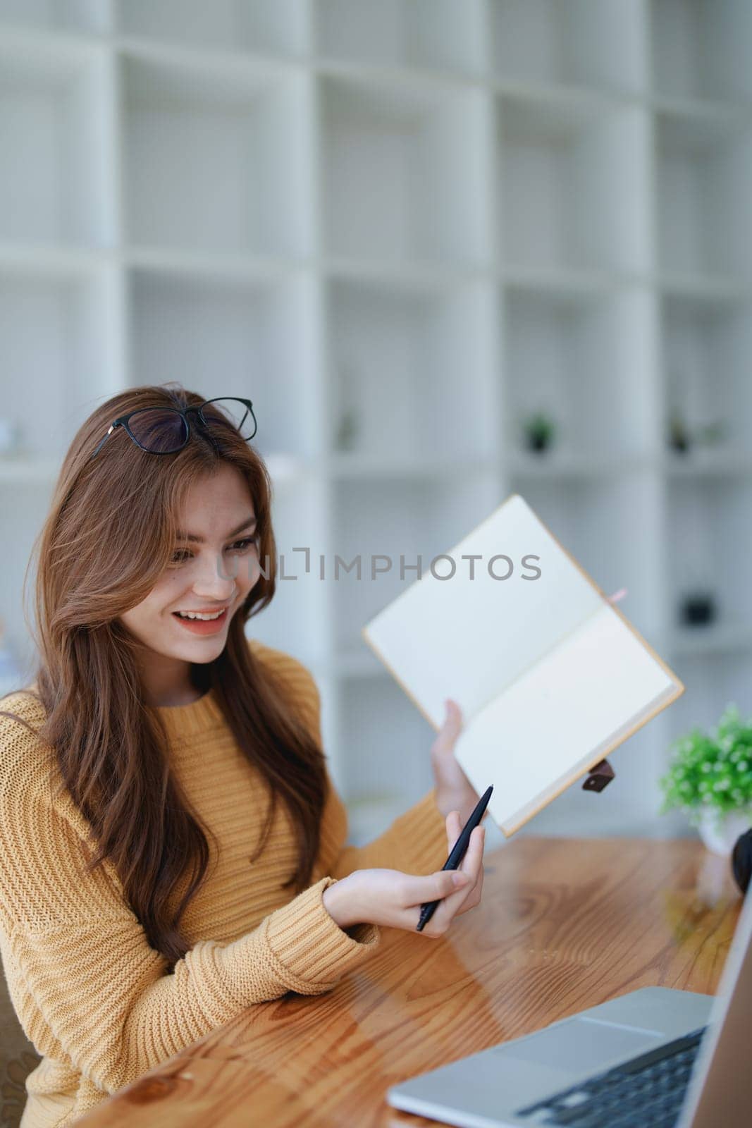 Smiling young woman engaged in a joyful online e-learning video call, gesturing while holding a book with headphones on a wooden table nearby.