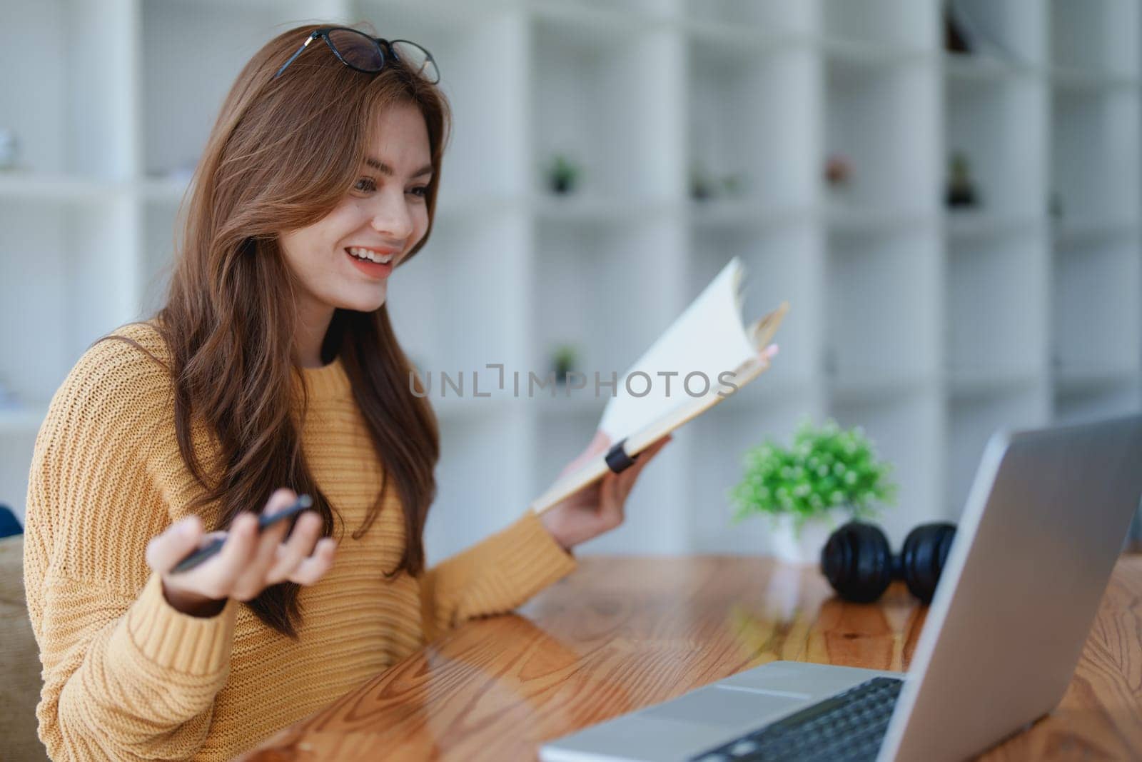 Smiling young woman engaged in a joyful online e-learning video call, gesturing while holding a book with headphones on a wooden table nearby.