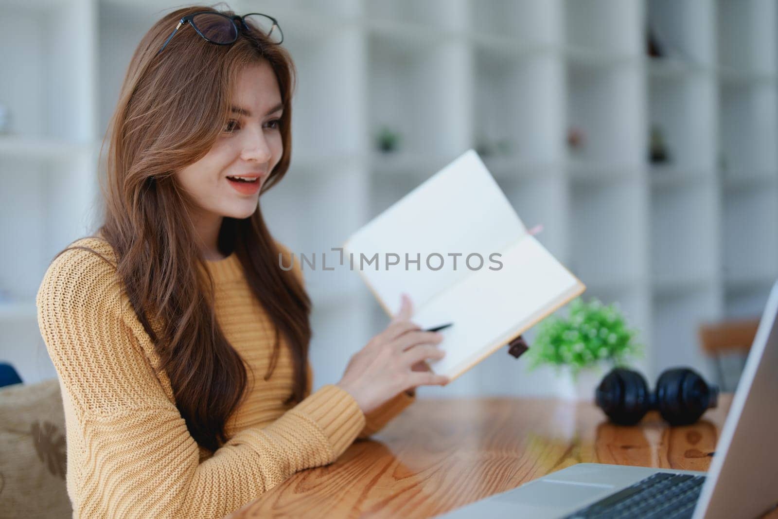 Smiling young woman engaged in a joyful online e-learning video call, gesturing while holding a book with headphones on a wooden table nearby.