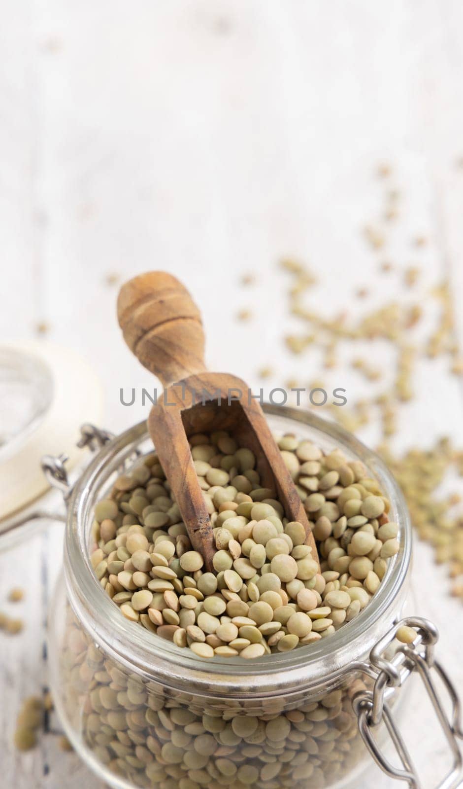 Opened glass jar full of dried green lentils with a wooden scoop on white table closeup. Healthy vegetarian food, legumes plant-based protein source. Traditional mediterranean raw uncooked ingredient