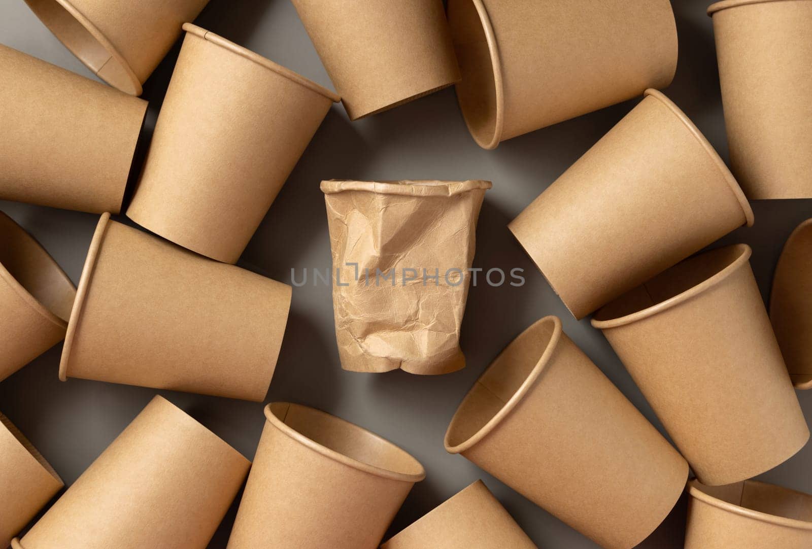 Disposable new beige kraftr paper cups and crumpled used one in middle on gray top view. Empty cardboard single-use coffee cups flat lay. Consumerism concept in modern world