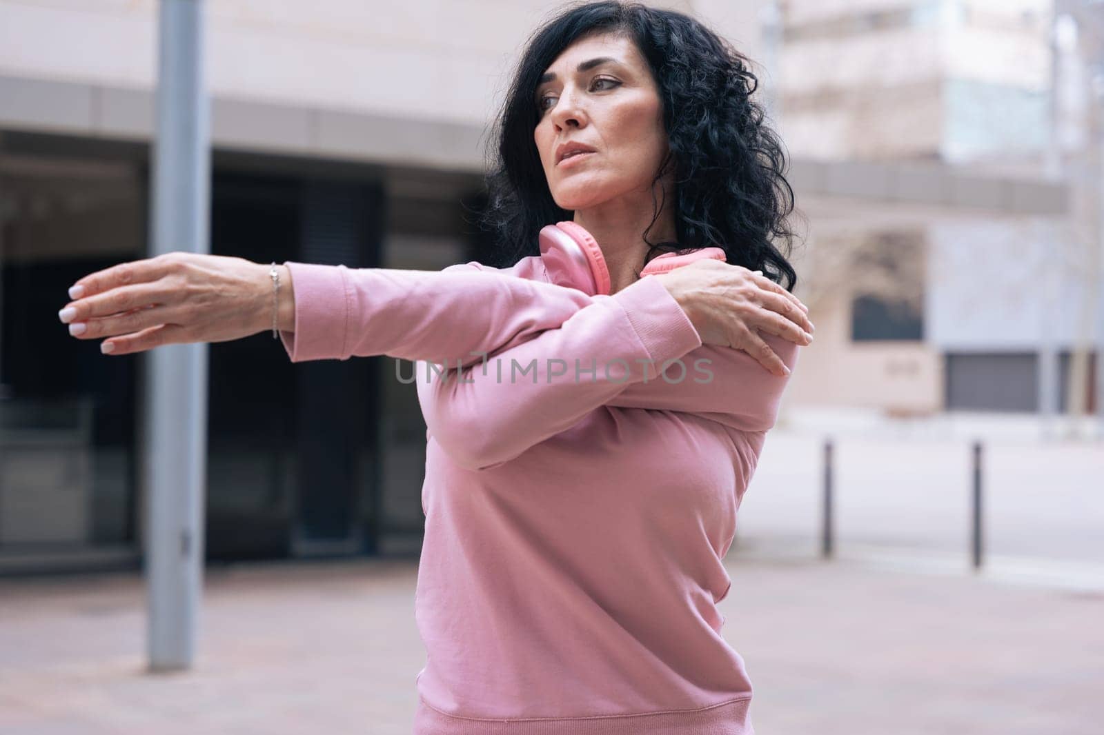Caucasian adult woman with curly black hair stretching her arms outdoors in an urban setting.she is wearing a pink sweatshirt and exercising for a healthy lifestyle, fitness, and wellness in the city