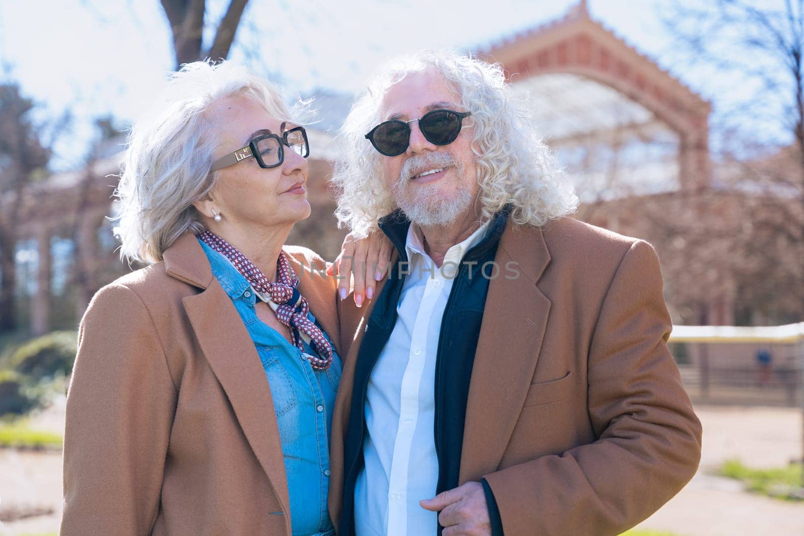 Caucasian elderly couple standing outdoors in a sunny park, smiling and posing together. The woman rests her arm on the man's shoulder while they enjoy a pleasant moment in the city.