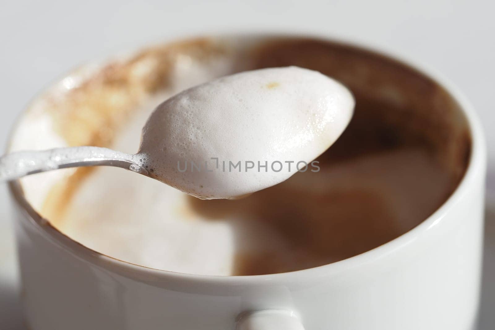 A spoon with rich foam hovers above a warm coffee cup on a table.