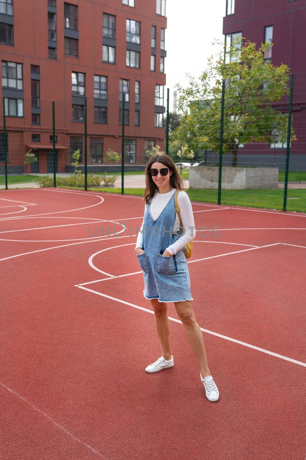 A young woman in a denim dress stands on a red outdoor basketball court, smiling confidently.
