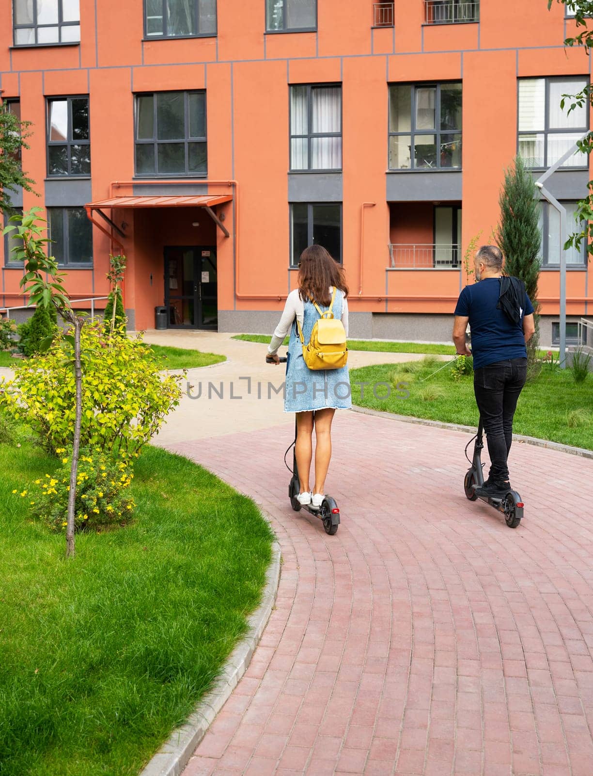 Two young adults enjoy a leisurely ride on electric scooters through a modern apartment complex's courtyard.