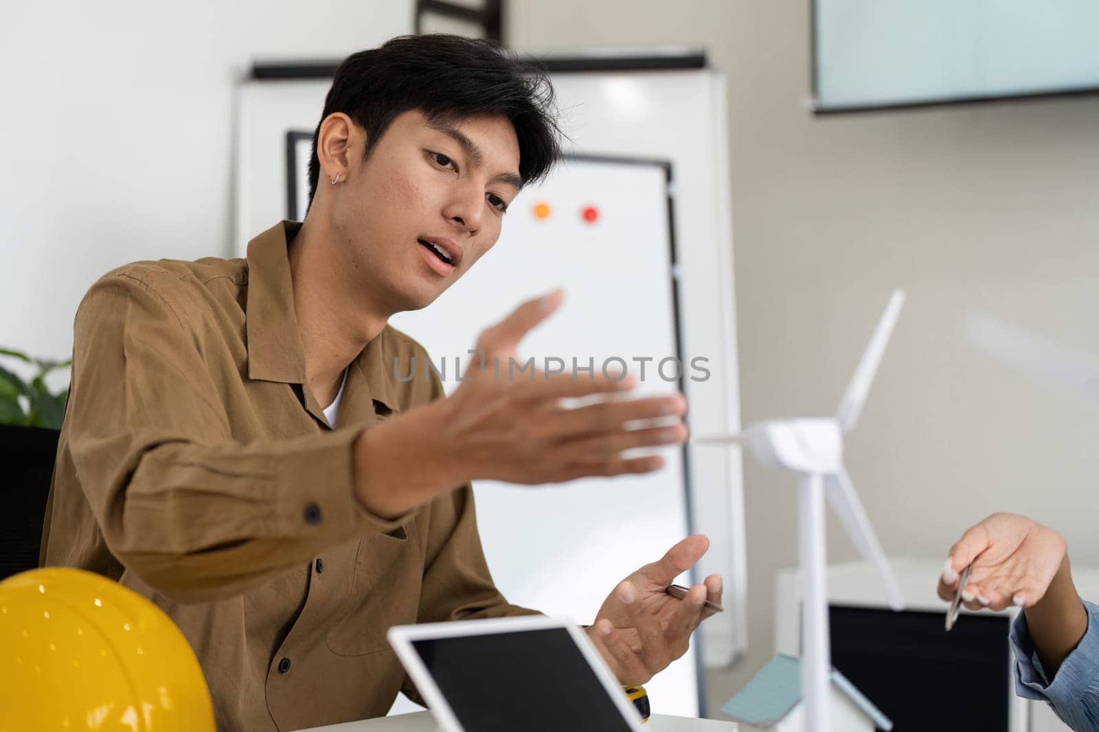 In a collaborative workspace, a young professional explains the benefits of wind energy while pointing at a miniature wind turbine model, signaling innovation in sustainable design.