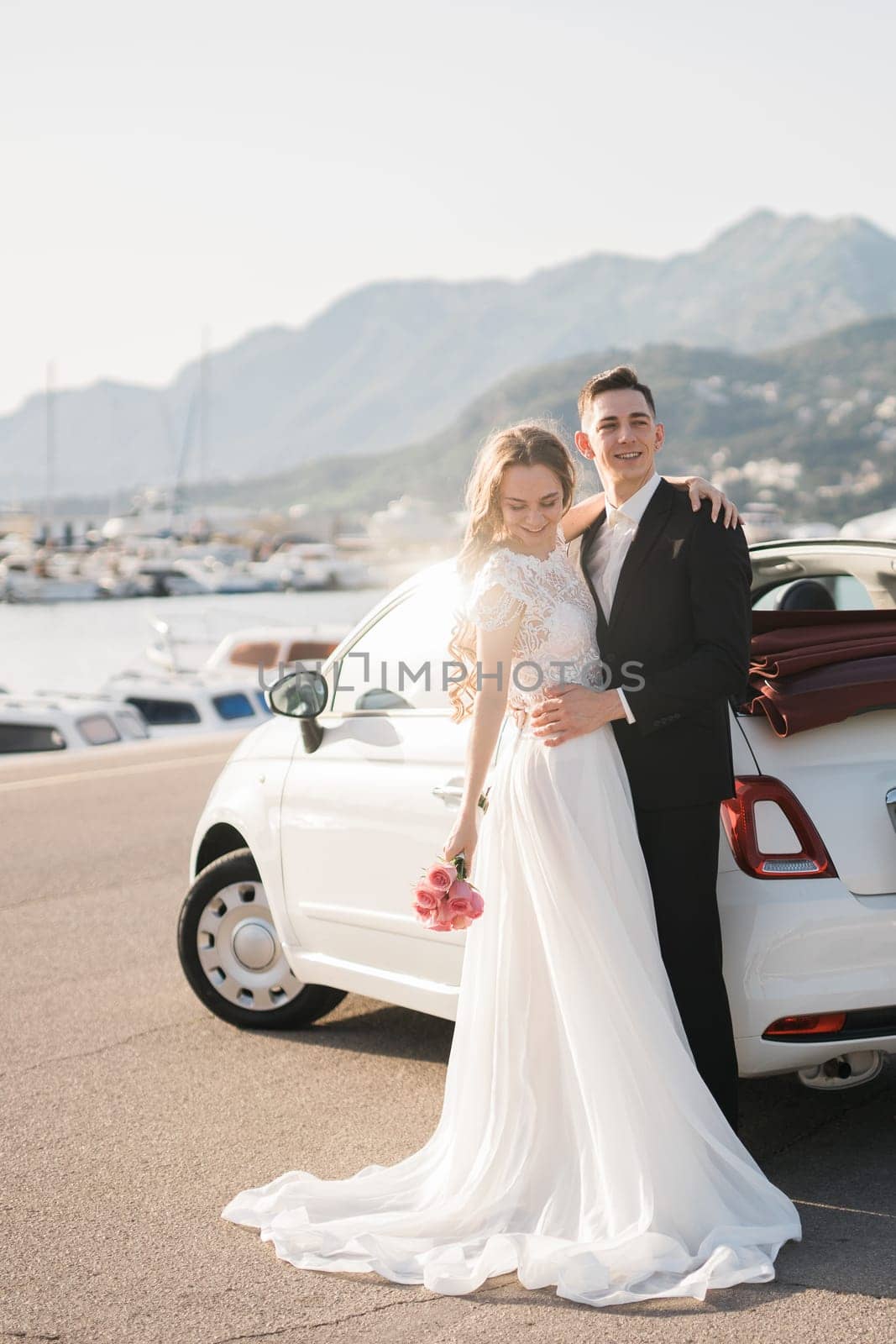 Stylish newlyweds are hugging near a white car in a green park, against the backdrop of summer nature. Bearded groom and blond bride in a lace dress are smiling outdoors. Wedding portrait.