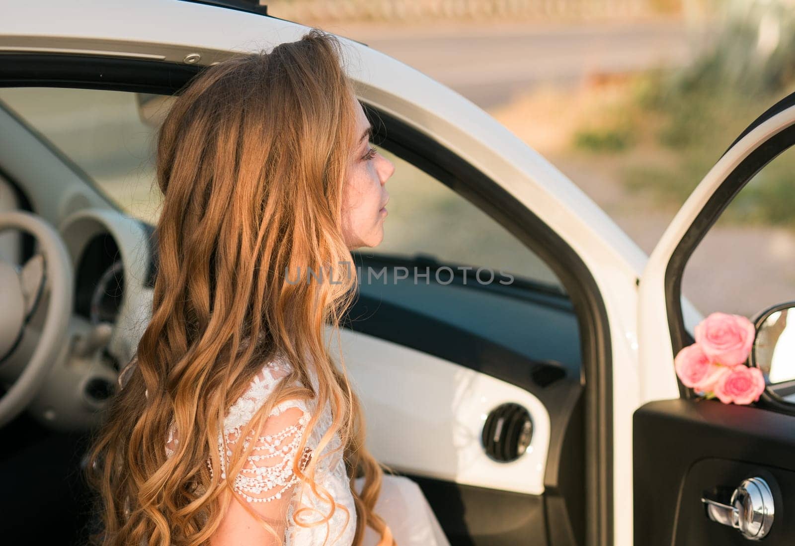 Young woman with long wavy hair sits at open car door. Femininity, romantic mood, travel moment and natural beauty in golden sunlight.