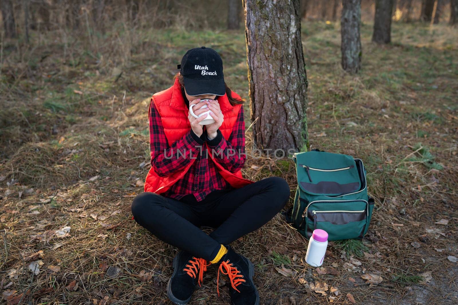 Trekking, hiking. A backpack with a thermos and a cup stands near a tree, a girl warms herself with hot tea, a snack in the open air.Travel and tourism