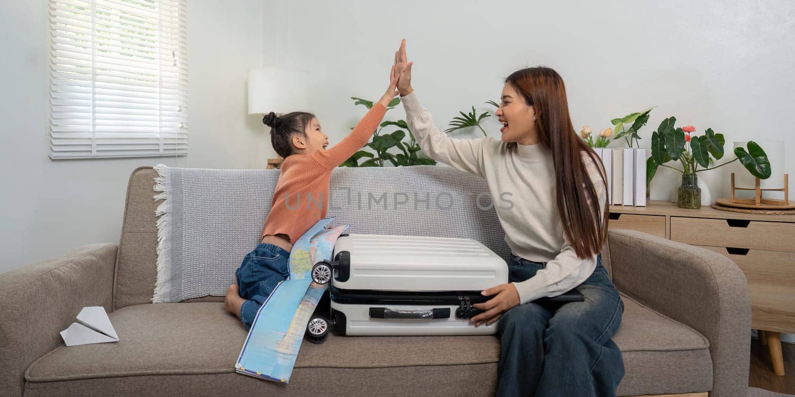 In a cozy living room, a mother and daughter enjoy a fun moment, high-fiving over their travel plans while sitting beside their suitcase.