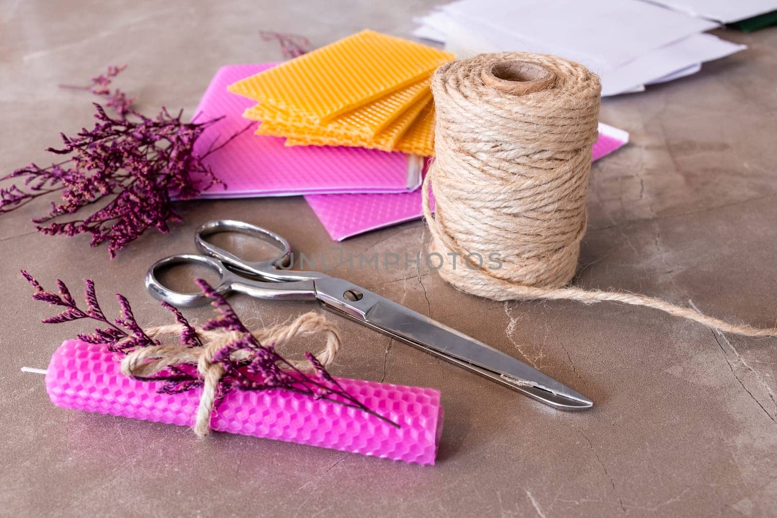 Crafting materials. Pink beeswax candle, twine, scissors, and purple flowers on concrete surface.