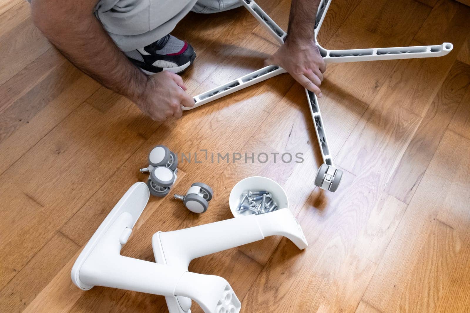 Adult caucasian male assembling office chair on wooden floor with tools and parts.