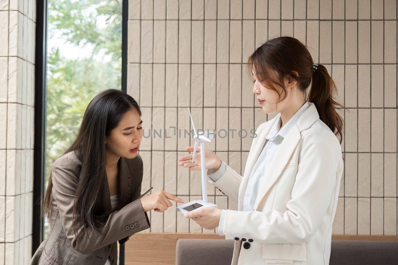 In a sleek workplace, two women engage in a conversation about sustainable technology, showcasing a miniature wind turbine model as they strategize advancements in ESG initiatives.