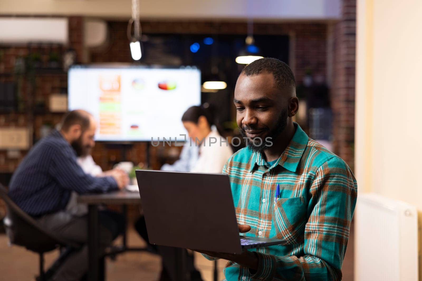 Professional black man deeply engaged in project planning, reviews financial analytics on his laptop. Behind him, coworkers discuss business trends, creating a dynamic workplace scene.