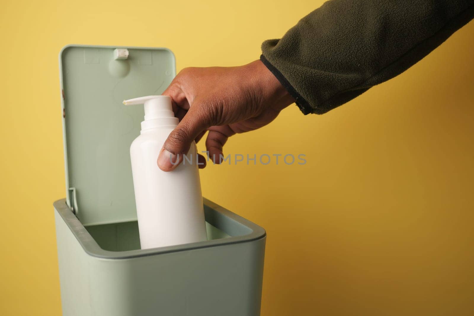 A hand drops a white bottle into a green trash can on yellow.