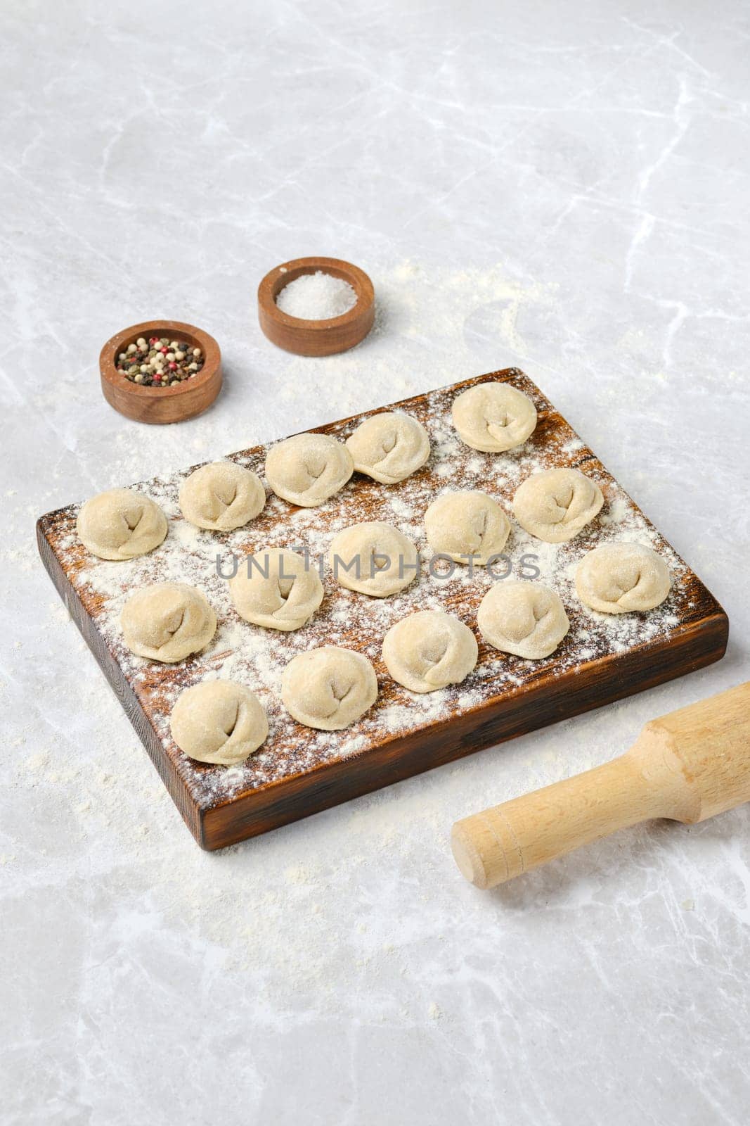 Dough dumplings with beef on a wooden board, dusted with flour. A rolling pin and bowls of spices surround the dumplings, hinting at a homemade cooking experience in a kitchen setting.