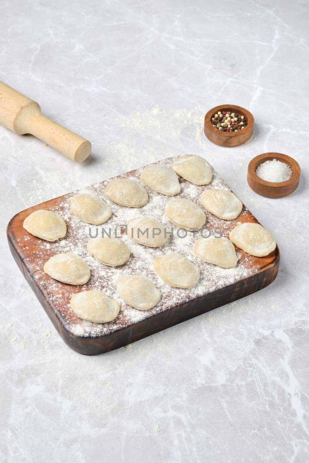Homemade dumplings are neatly arranged on a floured wooden board. Nearby, a rolling pin and small bowls of salt and pepper add to the cooking preparation atmosphere.