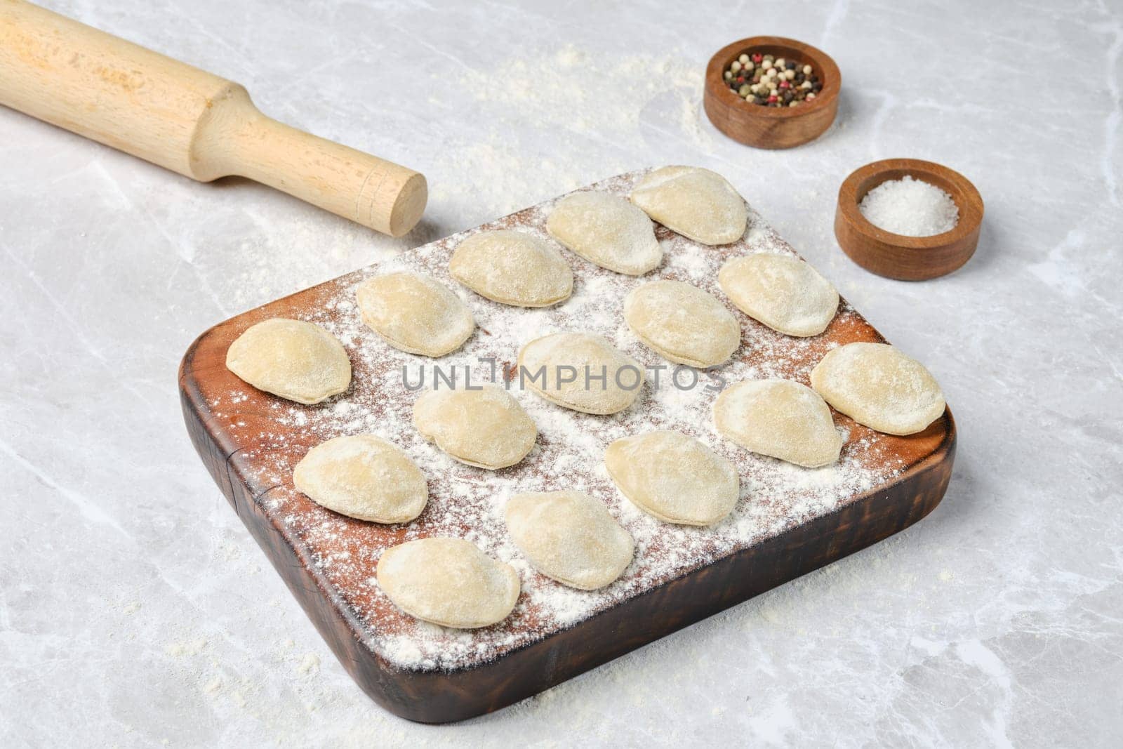 Dough dumplings with meat on a wooden board dusted with flour. A rolling pin rests nearby, indicating the process of preparing these tasty bites. Seasoning containers are visible.