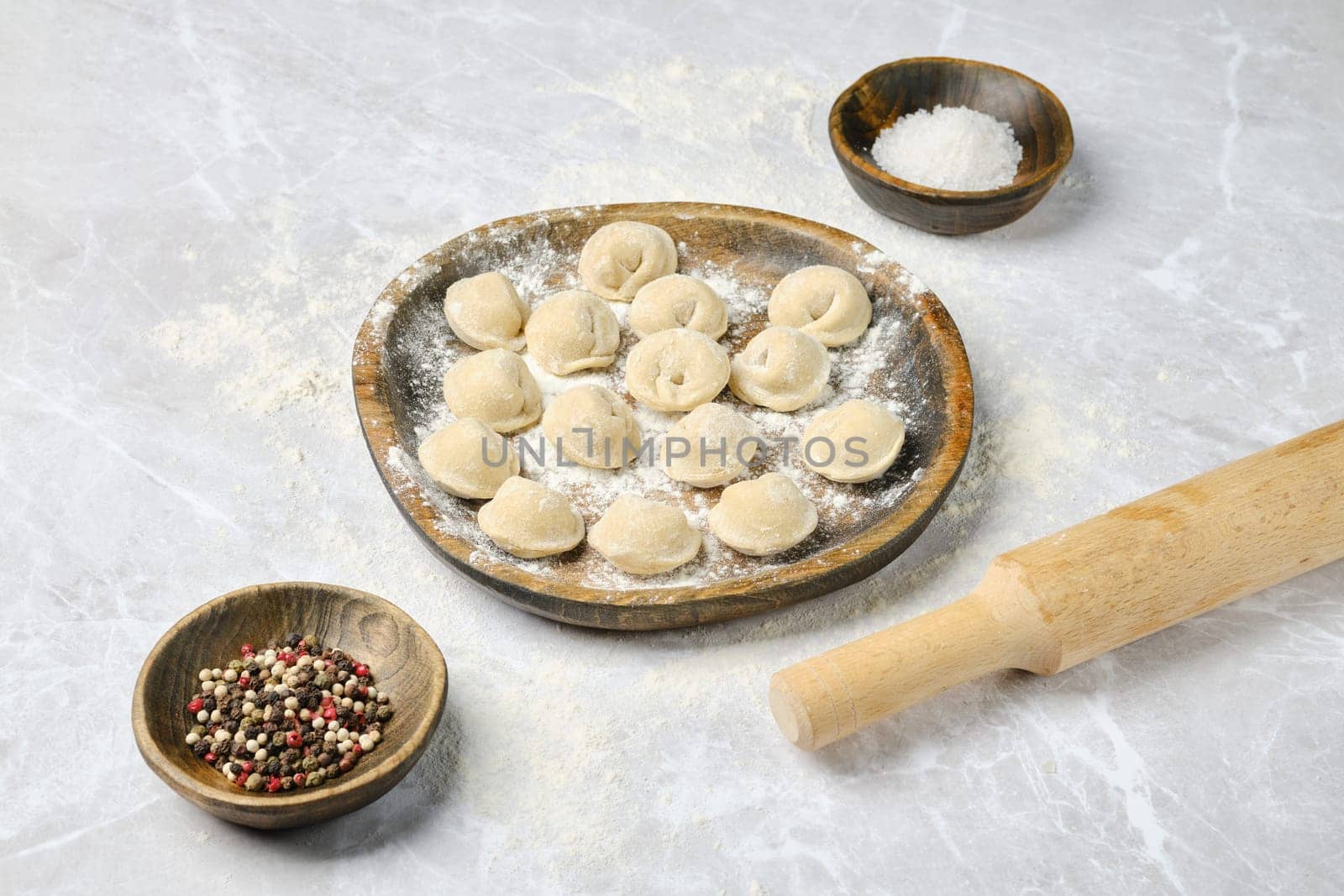 A wooden plate holds freshly made dumplings surrounded by bowls of spices and salt on a flour-dusted countertop. A rolling pin rests nearby, indicating the process of making these treats.