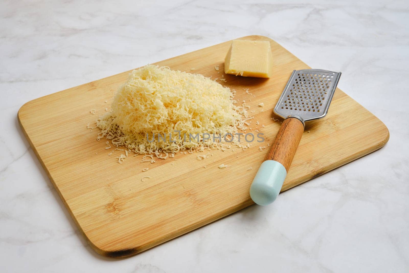 A wooden cutting board holds a pile of finely shredded cheese beside a block of cheese and a metal grater. The items are arranged neatly, showcasing a kitchen preparation setup.