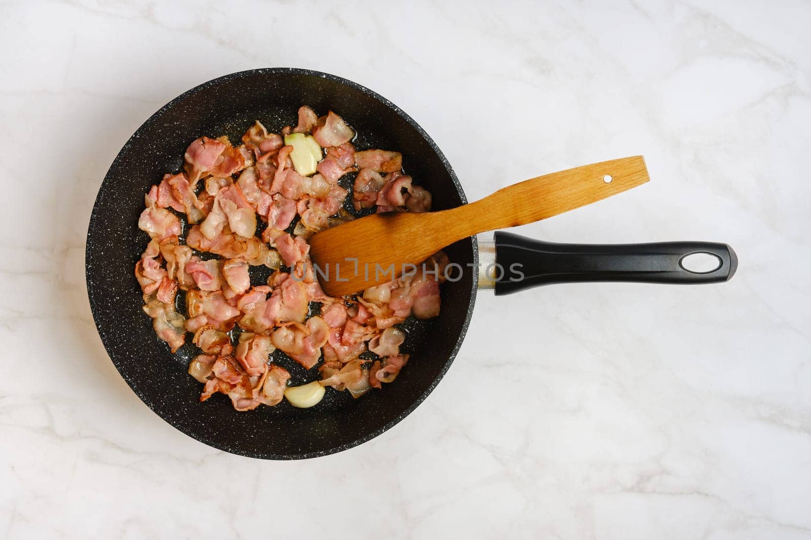 Bacon in a black frying pan on a marble countertop. The crispy pieces of bacon are accompanied by a wooden spatula, showcasing a delicious and appetizing preparation.