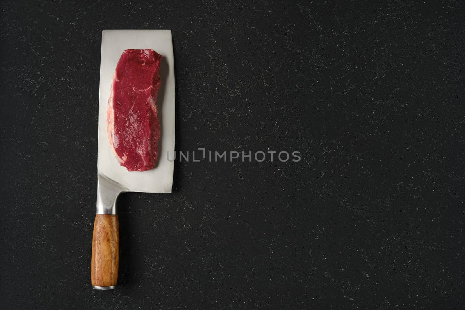 A piece of raw beef meat on a sharp cleaver with a wooden handle. The cleaver rests on a black countertop, showcasing the texture of the meat and the gleam of the blade.