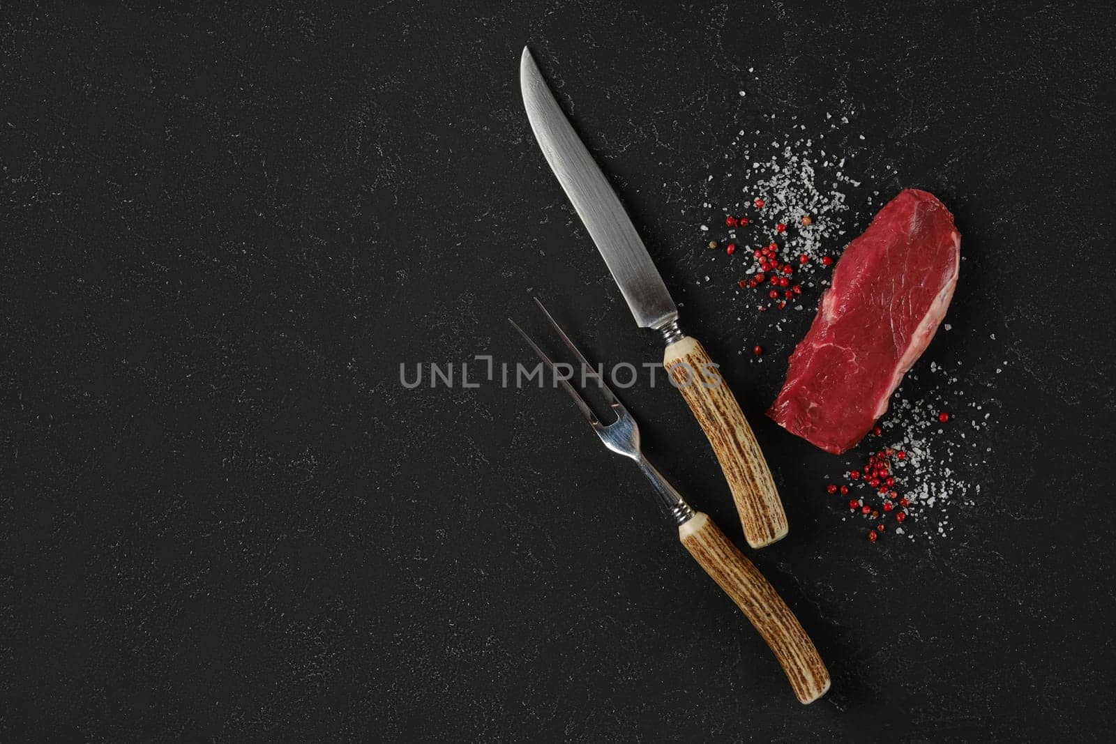 A knife and fork, with horn handles, rest beside a raw beef steak on a dark surface. Salt and pink peppercorn surround the meat, suggesting preparation for cooking or a gourmet meal.